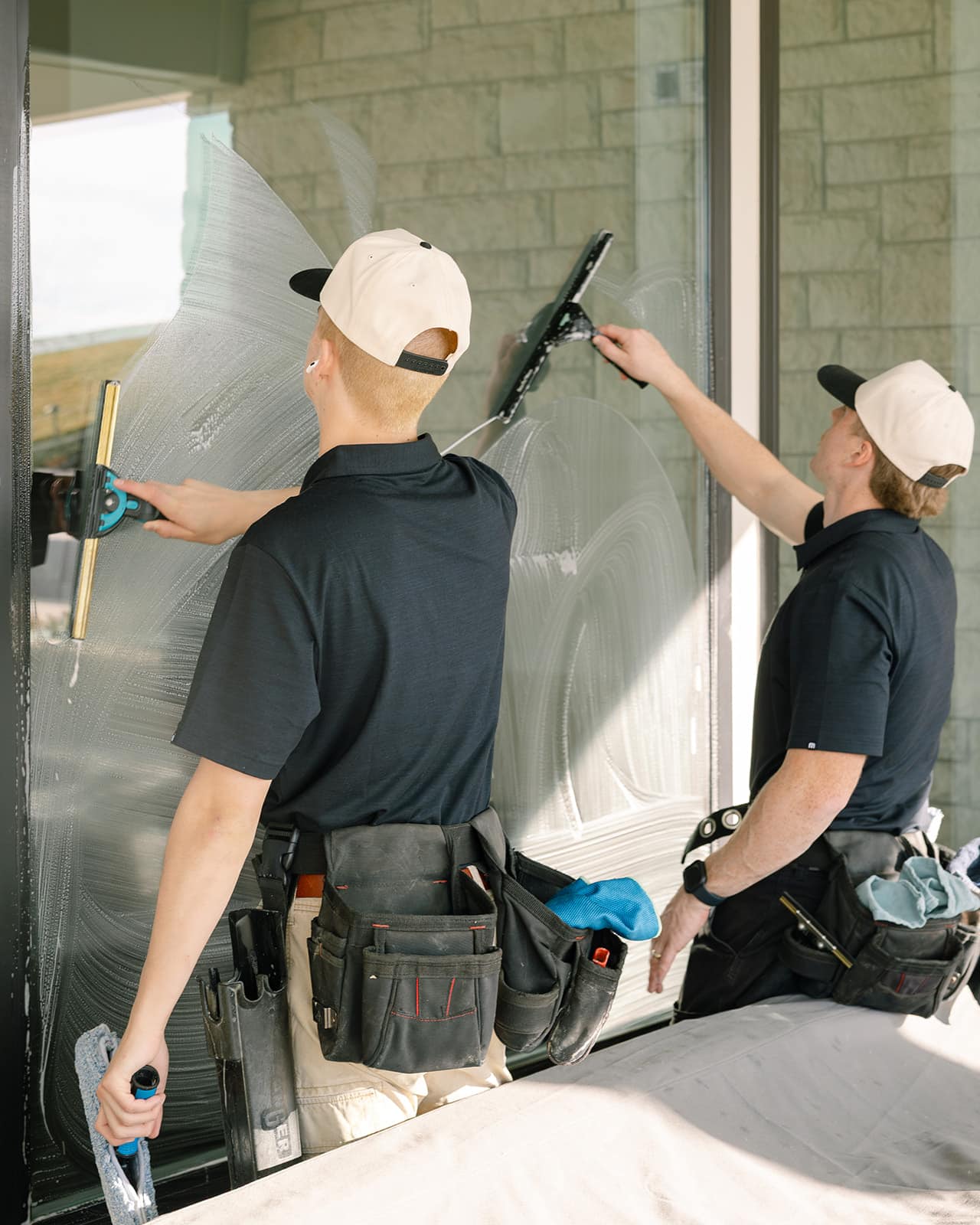 Two professional window cleaners from Bishop Window Cleaning in Logan, Utah, are washing large residential windows with squeegees.