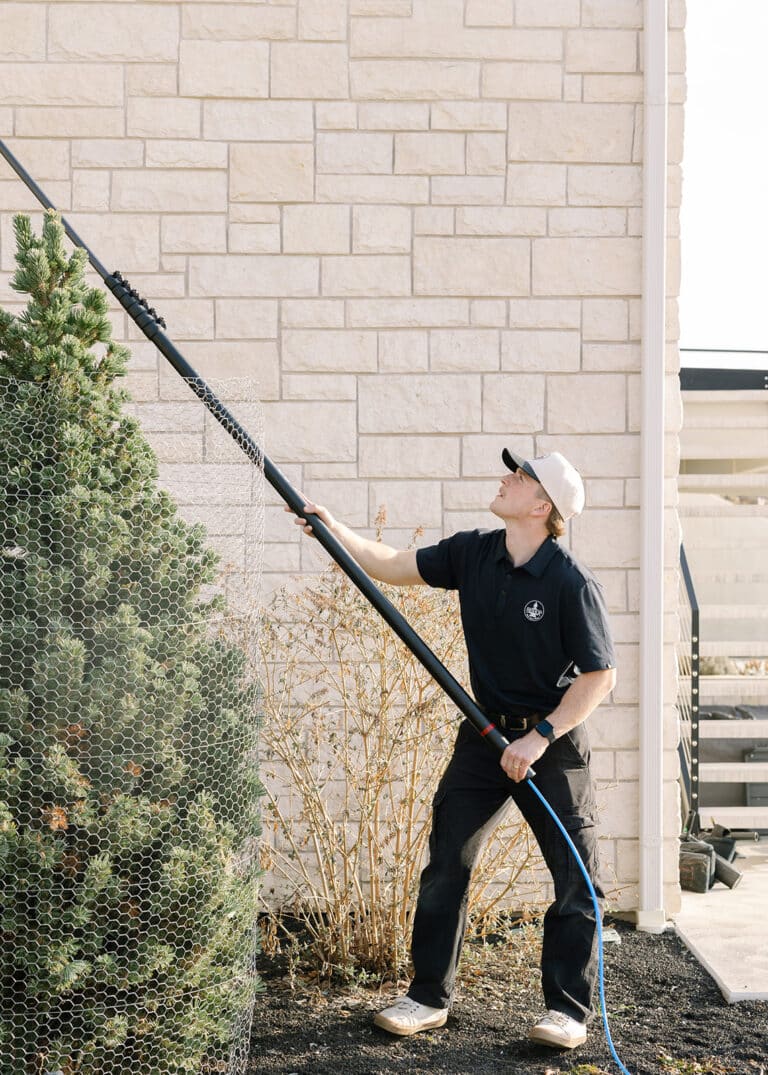 A Bishop Window Cleaning technician uses a water-fed pole to clean windows on a light brick house in Logan, Utah.