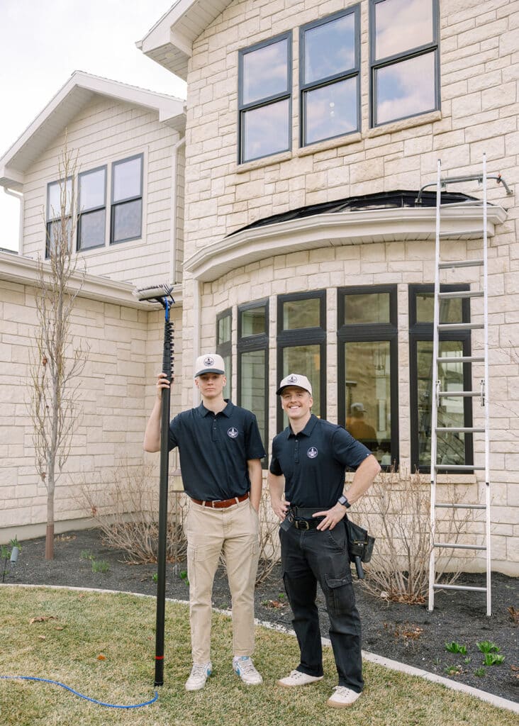 Two professional window cleaners in branded uniforms stand before a large stone home in Logan, Utah, holding a water-fed pole and ladder.