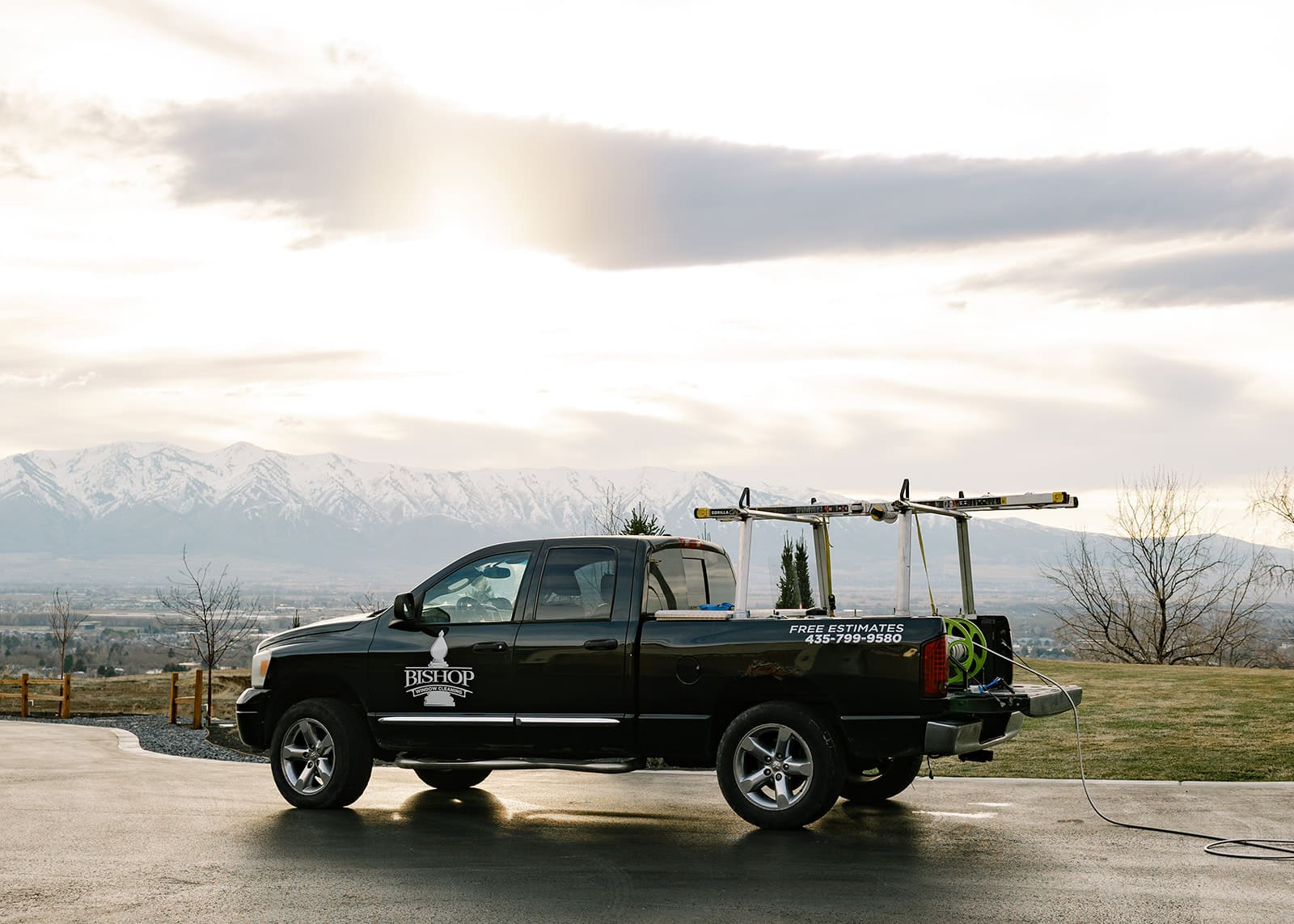 Black Bishop Window Cleaning truck with ladders and hose reel, parked in Logan, Utah, with snow-capped mountains in the background. Offers free estimates.